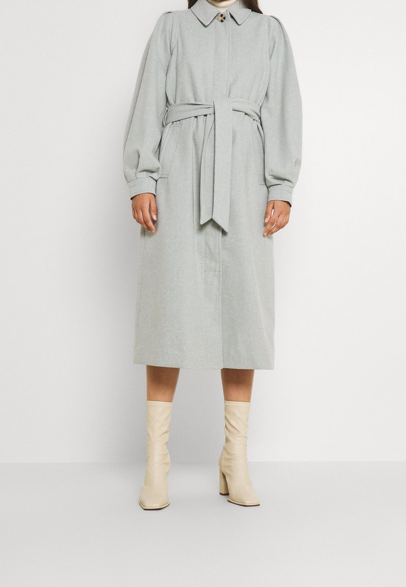 Manteau gris clair, longueur genou, avec une taille ceinturée, un grand col et des manches volumineuses. Associé à des bottes stiletto crème jusqu'au genou.