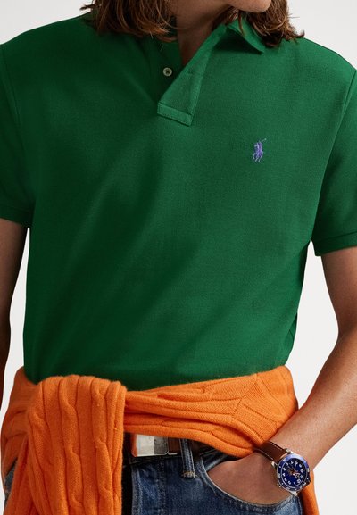 Πράσινο πόλο πουκάμισο με μοβ λογότυπο, σε συνδυασμό με πορτοκαλί πλεκτό πουλόβερ δεμένο γύρω από τη μέση. Φαίνονται τζιν παντελόνι και μπλε ρολόι.