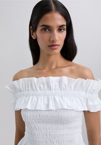Ung kvinna med långt svart hår bär en vit off-shoulder klänning med ryschkrage och smockad midja mot en enkel ljus bakgrund.