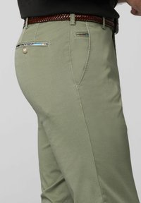 Groene broek met een gestructureerde afwerking, voorzien van een zijzak met decoratieve stiksels en een bruin gevlochten riem.