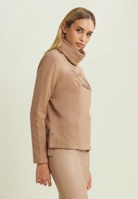 Felpa beige con colletto alto, caratterizzata da una grafica metallica sulla parte anteriore, abbinata a leggings attillati di colore chiaro.