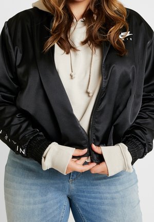 Personne portant une veste noire Nike Air, la dézippant sur un sweat à capuche beige avec des trous pour les pouces, associée à un jean bleu clair.
