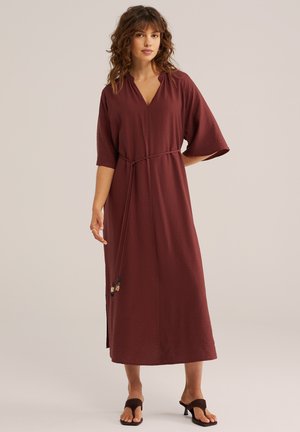 Frau trägt ein locker sitzendes, knöchellanges weinrotes Kleid mit halblangen Ärmeln und einem dünnen Gürtel, steht entspannt vor einem schlichten Hintergrund.