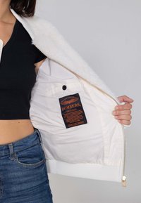 Witte bomberjack met ritssluiting, zachte textuur en geribde manchetten. Aan de binnenkant bevindt zich een merklabel en een knoopzak.