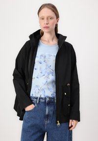 Μαύρο αδιάβροχο μπουφάν με κουκούλα, κλείσιμο με φερμουάρ, δύο μπροστινές τσέπες. Φορεμένο πάνω από ένα ανοιχτό μπλε πουκάμισο με λουλουδάτο σχέδιο και ψηλόμεσα τζιν.