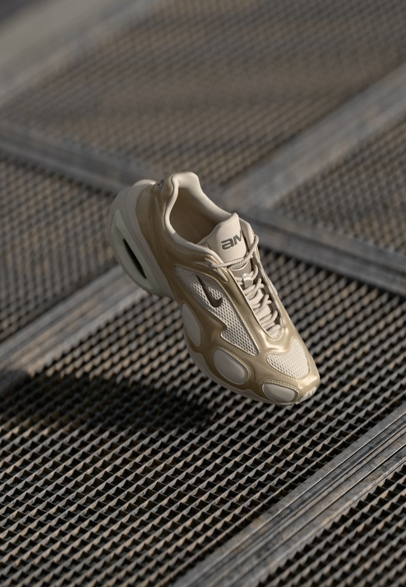 Shoe atlético bege com detalhes em malha e borracha, apresentando um design arredondado, sola proeminente e sistema de atacadores. Destacado sobre uma superfície texturizada.