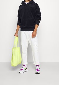 Schwarzer Hoodie mit einer Fronttasche, weiße Cargo-Hosen und weiße Sneaker mit lila und grünen Akzenten. Hält eine neongelbe Tragetasche.
