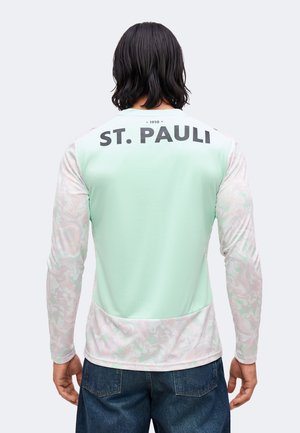 Personne aux cheveux foncés mi-longs porte une chemise à manches longues vert clair à motifs avec le texte « ST. PAULI » en haut du dos.