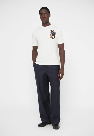 T-shirt en coton blanc avec un motif coloré de chat sur la poitrine gauche, assorti à un pantalon à fines rayures bleu marine et des chaussures noires.