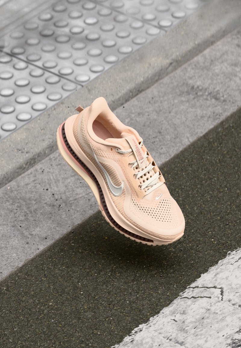 Chaussure de course Nike rose clair suspendue au-dessus d'un trottoir urbain texturé avec un revêtement tactile et de l'asphalte visible.
