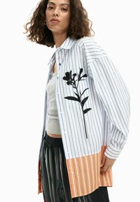 Sinine ja valge triibuline nööbitav särk, mille ühel küljel must lillemuster, ere oranž alläär ja lõdvestunud istuvus.