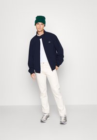 Mornarska zip-up jakna, bela majica, smetana hlače, sivi športni copati in zeleni zimski klobuk. Preprost dizajn, sproščen kroj in mehak material.