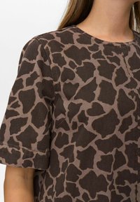 Hnědý krátký rukáv top s designem ve stylu žirafy, kde tmavší skvrny jsou na světlém podkladu. Materiál má texturovaný povrch.