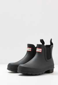 Bottes de cheville en caoutchouc noir avec des panneaux latéraux élastiques, une semelle rainurée et une étiquette logo rouge et blanche sur le haut de la tige. Texture lisse.