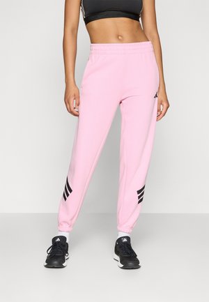 Woman wearing light pink Adidas joggers with black logo stripes near ankles, black sports bra, white socks, and black Adidas sneakers.