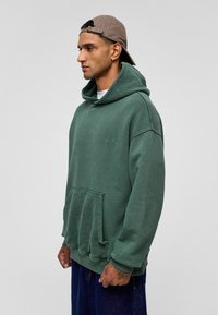 Groene hoodie van zacht materiaal, met een voorzak in de vorm van een kangoeroe en ribgebreide manchetten. Draagt een beige pet.