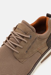 Sneakers en maille et en daim marron avec lacets beiges, semelle en caoutchouc et subtils accents orange. Logo sur le côté et matériaux texturés.