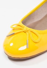Ballerine jaune brillante avec nœud tressé et bouts métalliques, doublure intérieure beige et semelle lisse sur fond blanc.