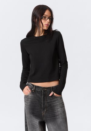 Femme aux longs cheveux noirs et lunettes, portant un crop top noir à manches longues et un jean noir délavé, les mains dans les poches.