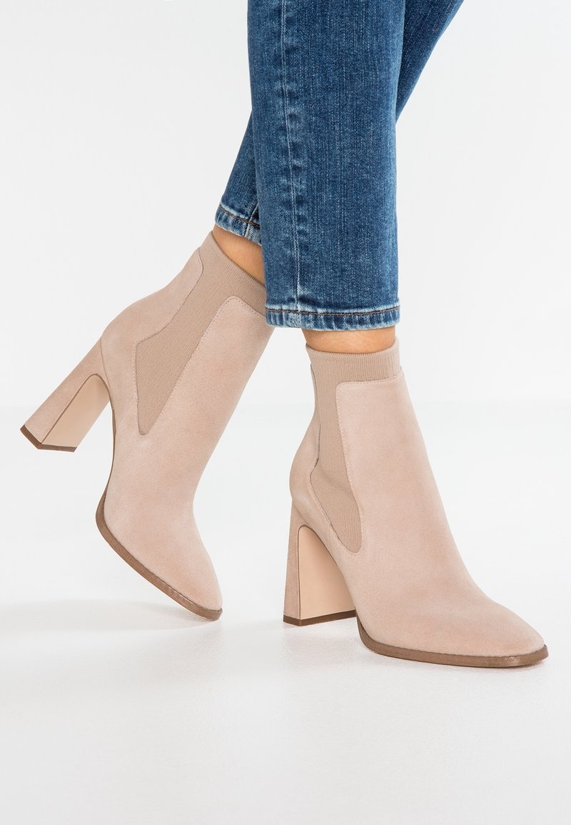 Botines de ante beige con paneles laterales elásticos y tacón grueso. Diseño de punta puntiaguda, textura suave, y detalles en crema a contraste.