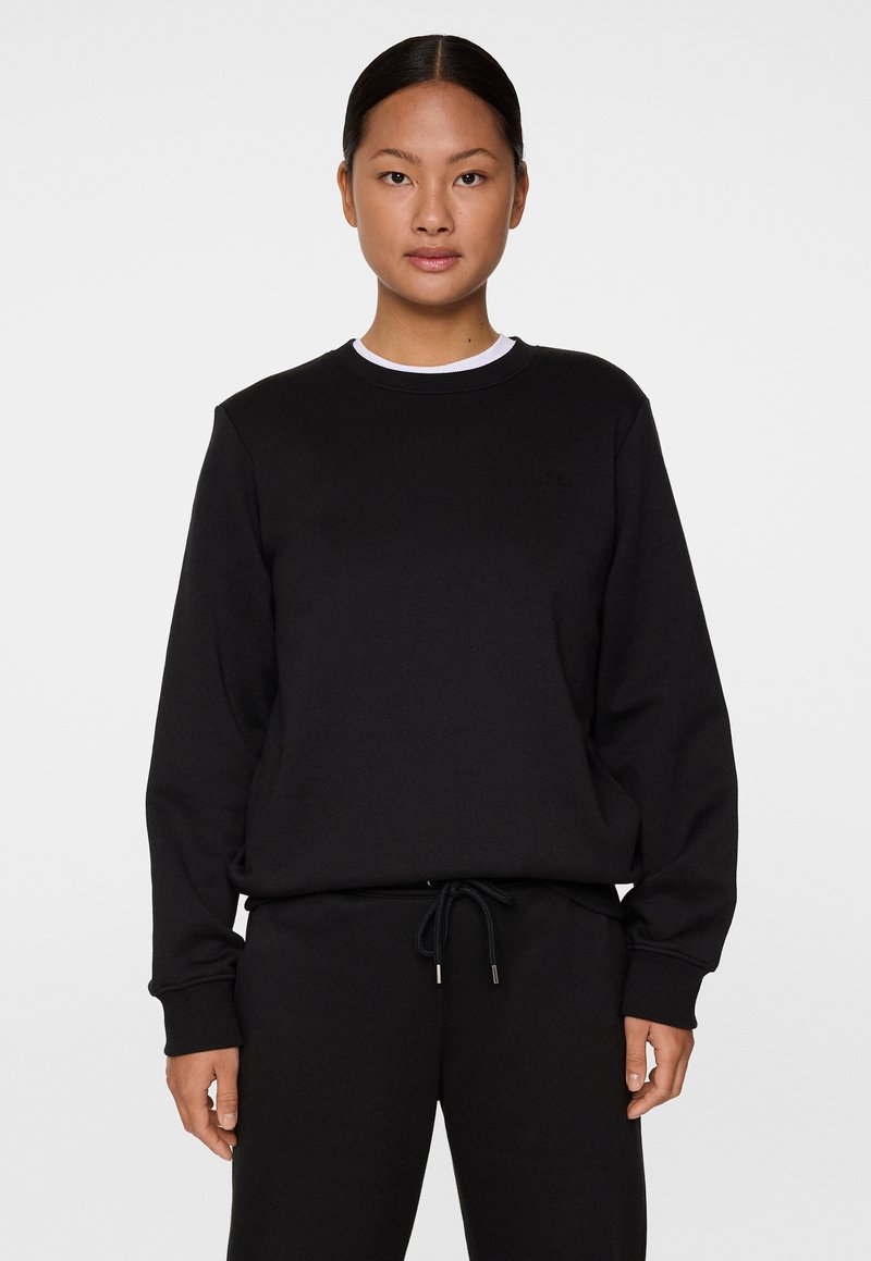 Schwarzes Sweatshirt aus weichem Material mit rundem Halsausschnitt, gerippten Bündchen und einem dezenten Logo-Detail. Kombiniert mit passenden schwarzen Hosen.