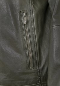 Dunkelgrüner Lederjackenabschnitt mit einer Reißverschlusstasche, die einen metallischen Reißverschluss und eine glatte Oberflächenstruktur aufweist. Sichtbare Nähdetails.