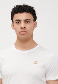 Biela krátkopesťová tričko z jemnej tkaniny, s malým zlatým vyšitým logom na ľavej strane hrudníka. Okrúhly výstrih.