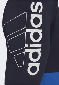 Leggings navy avec un grand logo "adidas" blanc et un motif géométrique. Fabriqués en tissu extensible avec une texture lisse.