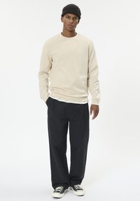 Krémovo béžová mikina s dlhými rukávmi, v kombinácii s voľnými čiernymi nohavicami a čiernymi teniskami. Šité detaily a rebrovaná textúra na čiapke.