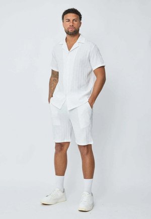 Chemise blanche à manches courtes avec un motif texturé, associée à un short blanc assorti avec des poches. Portée avec des baskets blanches et des chaussettes.