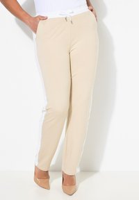 Béžové tepláky s bílým stahovacím pasem, bočními kapsami a bílými texturovanými akcenty podél bočních švů, kombinované s nahými podpatky.