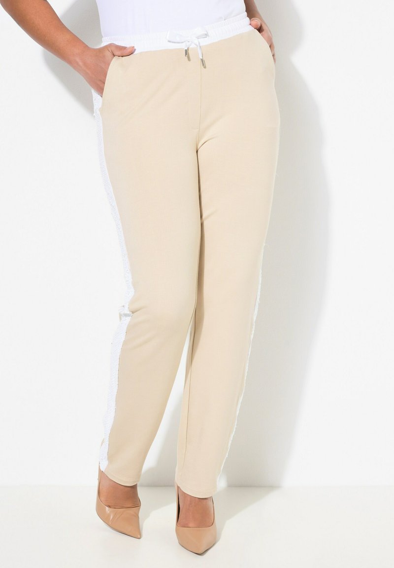 Béžové tepláky s bílým stahovacím pasem, bočními kapsami a bílými texturovanými akcenty podél bočních švů, kombinované s nahými podpatky.