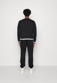 Sudadera de algodón negra con puños y cinturilla a rayas, combinada con joggers negros, vista desde atrás. Zapatillas blancas visibles.