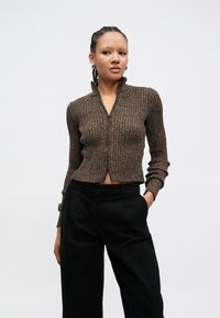 Cárdigan corto de punto acanalado marrón con cremallera frontal y cuello alto, combinado con pantalones negros de pierna ancha y cintura alta. Textura suave.