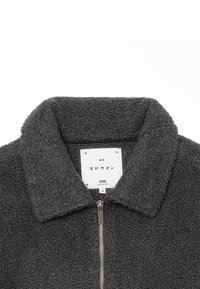 Veste polaire gris foncé avec texture, fermeture éclair à l'avant et une étiquette blanche indiquant le nom de la marque et la taille à l'intérieur du col.