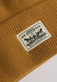 Mustard yellow knit beanie with ribbed texture. Features a rectangular Levi Strauss & Co. patch detailing horses and text.