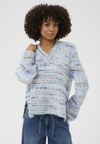 Femme jeune avec des cheveux bouclés portant un pull en maille multicolore bleu et gris ample, et un jean bleu, posant avec une main sur l'épaule.