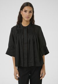 Black blouse with a high collar, featuring vertical pleats and textured panels. The sleeves are bell-shaped and made from lightweight fabric.
