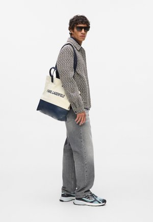 Medvilninis šoninis krepšys su tamsiai mėlynu dugnu ir rankenomis, grietinėlės spalvos kūnas, ant kurio užrašyta "KARL LAGERFELD". Modelis vilki raštuotą švarką ir pilkas džinsus.