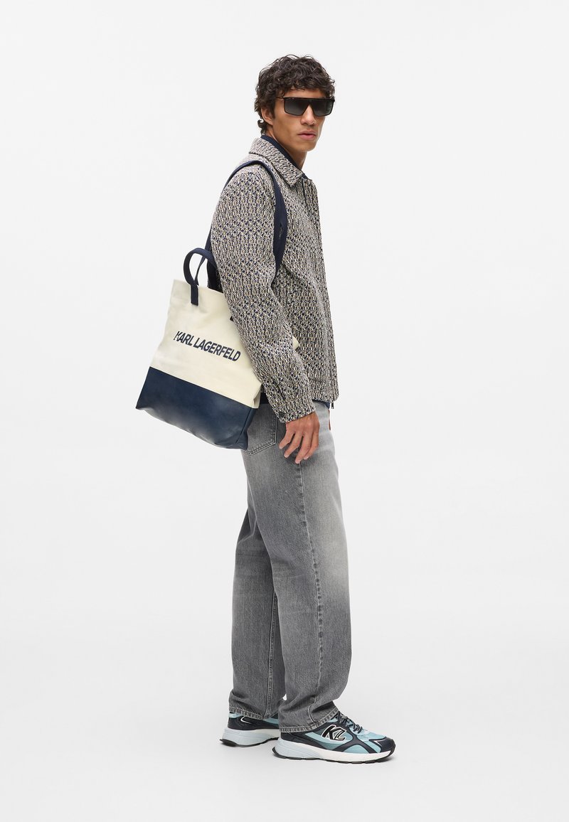 Medvilninis šoninis krepšys su tamsiai mėlynu dugnu ir rankenomis, grietinėlės spalvos kūnas, ant kurio užrašyta "KARL LAGERFELD". Modelis vilki raštuotą švarką ir pilkas džinsus.