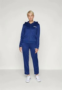 Melnā divdaļīgā sporta kostīms, kas izgatavots no mīksta auduma; kapuce ar rāvējslēdzēju un logo, atbilstošas bikses ar elastīgām aprocēm, kombinētas ar baltām sporta kurpēm.