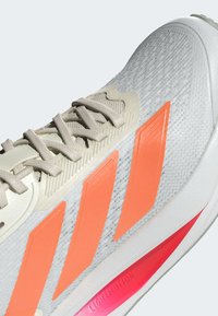 Zapatilla deportiva blanca con un upper texturizado, cordones beige y un diseño de tres rayas en naranja brillante; presenta un acento rosa en la suela.