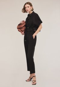 Zwart cropped shirt met schoudertabs, gecombineerd met zwarte op maat gemaakte broeken. Model houdt een gestructureerde rozenkleurige tas vast en draagt dunne zwarte sandalen.