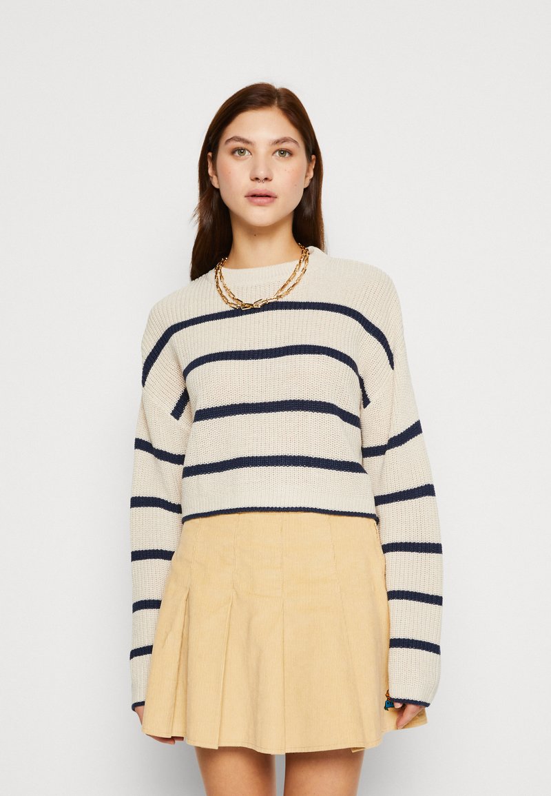 Pull en tricot beige avec des rayures horizontales bleu marine et un col rond, associé à une jupe en velours côtelé jaune clair plissée. Collier en chaîne dorée.