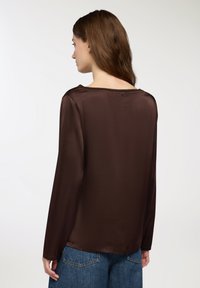 Blusa marrone a maniche lunghe con una texture liscia, scollatura ampia e dettagli di cuciture sottili, abbinata a pantaloni in denim.