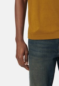 Maglione a maglia giallo senape con orlo a costine, abbinato a jeans in denim blu scuro. Primo piano di una mano appoggiata sui jeans.