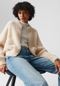 Junge Frau mit lockigem Haar, die eine beige, fuzzy Zip-Jacke, einen hellen, gepunkteten Rollkragenpullover und eine blaue Jeans trägt, sitzt auf einem schwarzen Hocker.