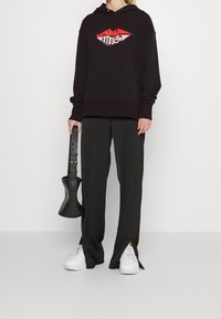 Czarna bluza z kapturem z graficznym wzorem w czerwone usta, w zestawieniu z czarnymi spodniami o szerokiej nogawce z bocznymi rozporkami oraz białymi sneakerami. Trzyma czarny przedmiot.