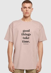 Lyserød oversized t-shirt lavet af bomuld, med sort tekst der siger "gode ting tager tid" og hashtagget "#TrustTheJourney."