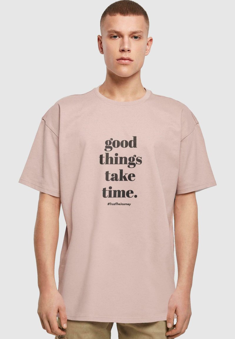 Lyserød oversized t-shirt lavet af bomuld, med sort tekst der siger "gode ting tager tid" og hashtagget "#TrustTheJourney."